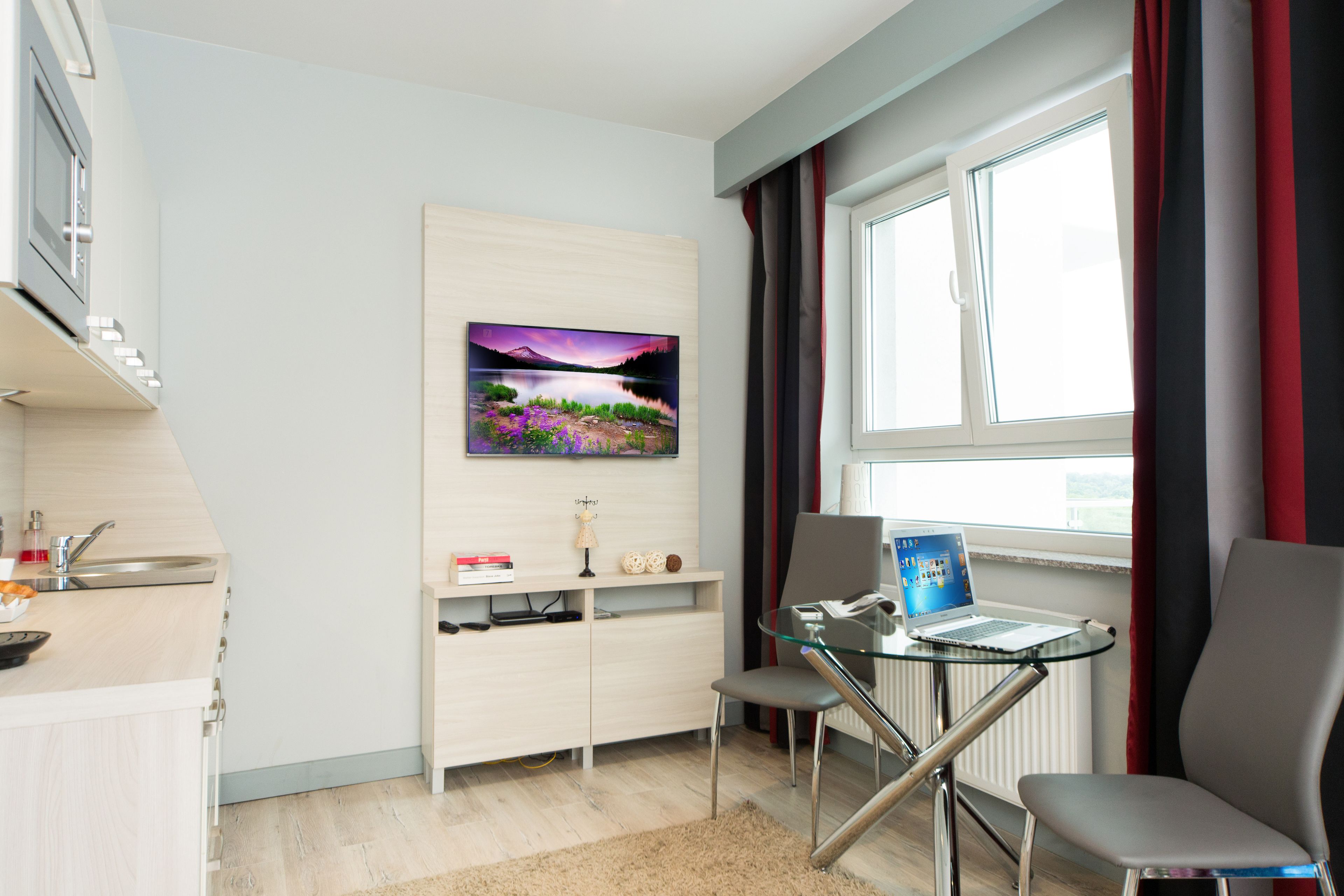 A modern kitchen with a built-in oven, a sink, and a countertop. There is a dining area with a glass table and two chairs. A painting is on the wall above the dining table.
