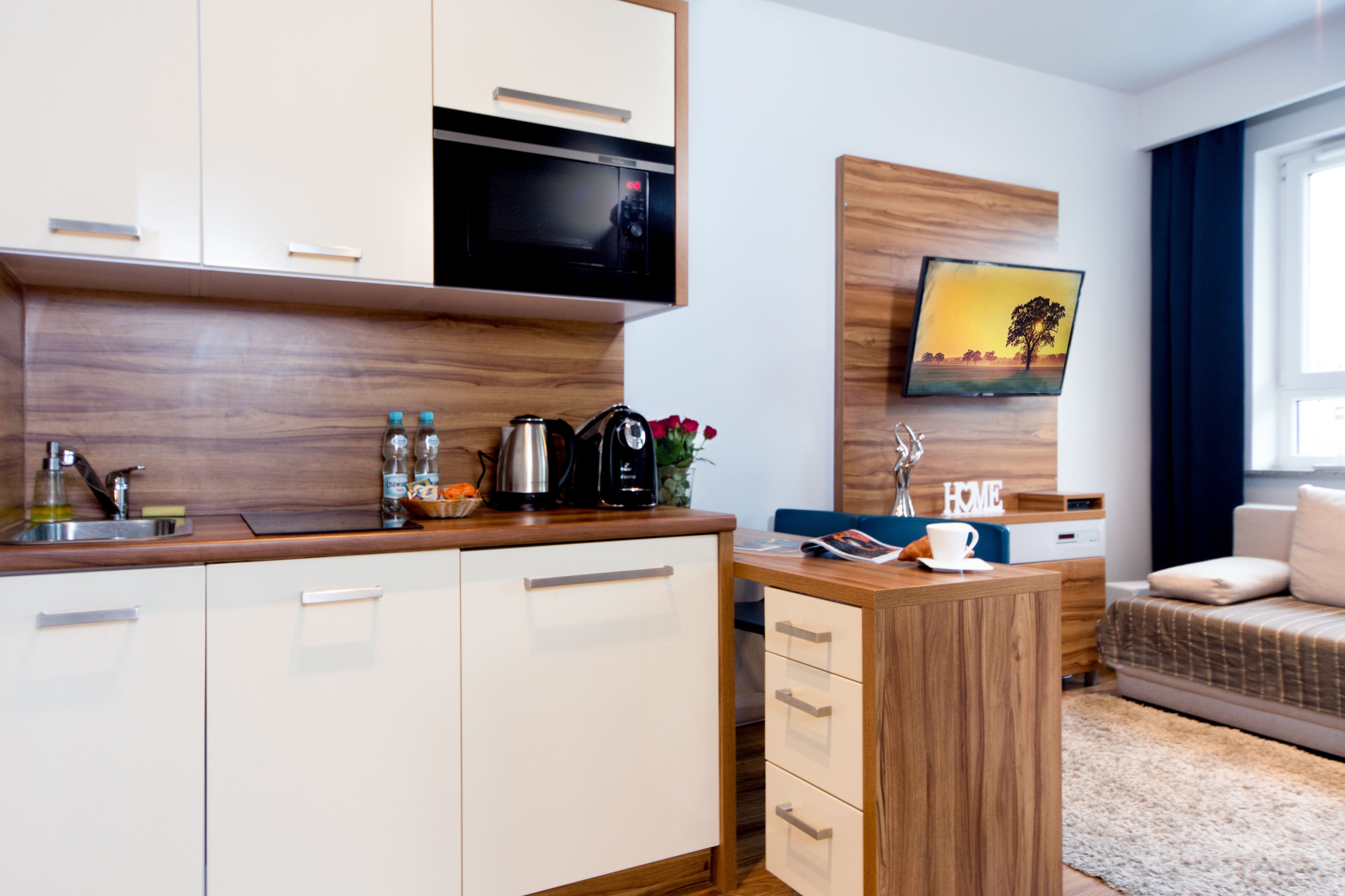 Una cocina moderna con microondas, cafetera y lavabo. Hay una encimera de madera y una pequeña zona de estar con sofá y mesa de centro. Un televisor está montado en la pared sobre la zona de estar.