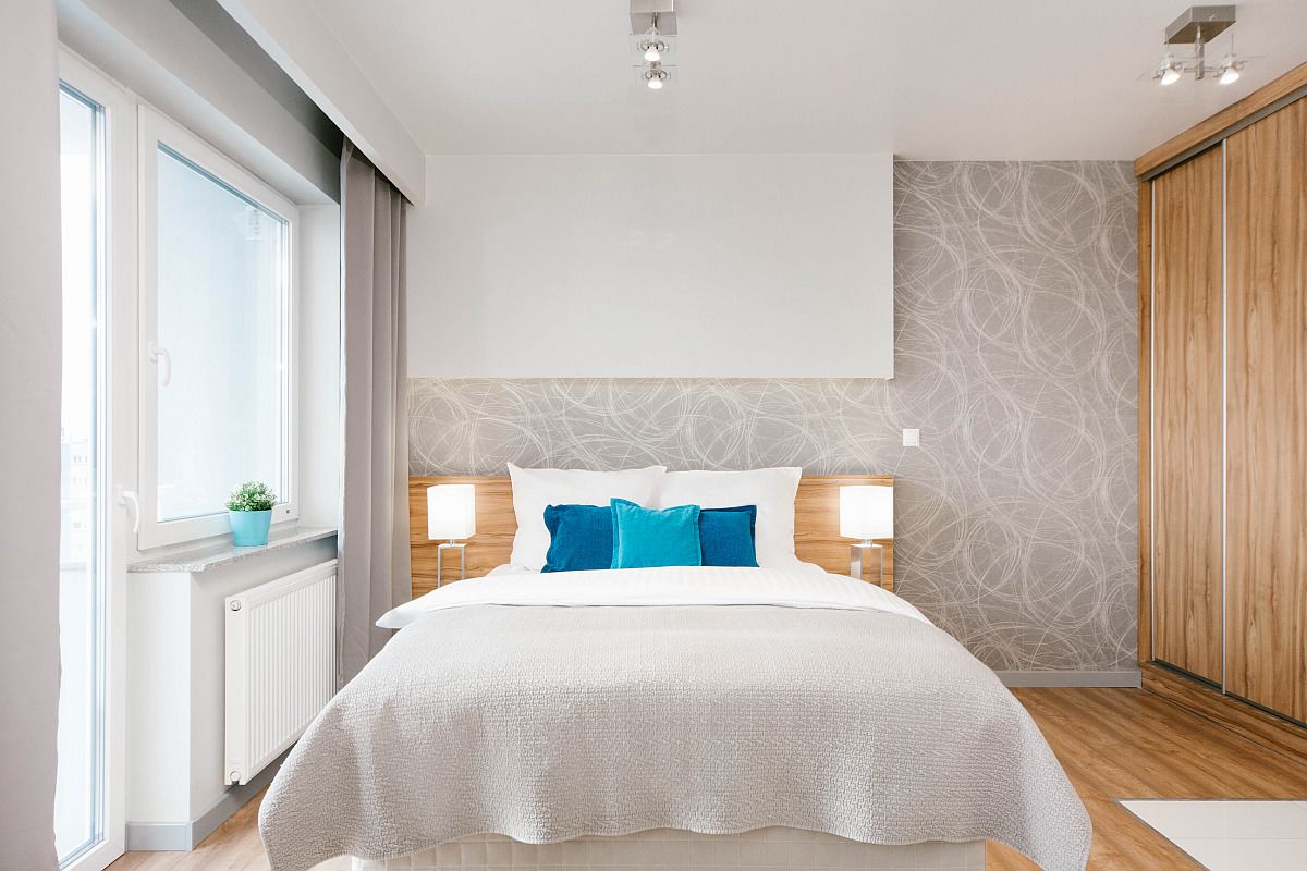 A modern bedroom with a large bed, two bedside lamps, a radiator, a window with curtains, and a patterned wall.