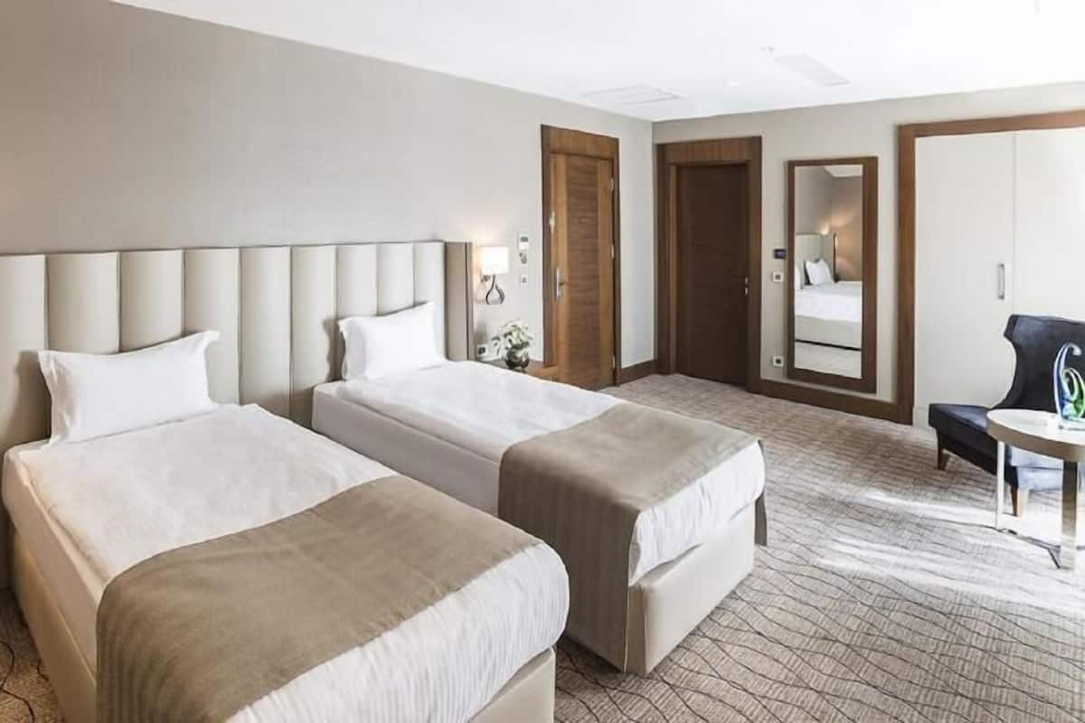 Quarto de hotel com duas camas, uma escrivaninha, uma cadeira, um abajur e um espelho.