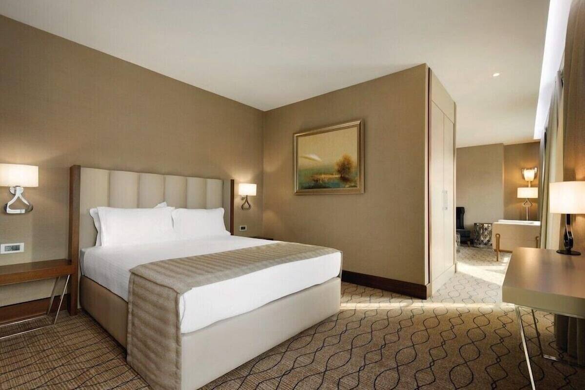 Quarto de hotel com uma cama grande, mesas de cabeceira, um quadro na parede e uma área de estar separada com uma mesa e cadeiras.