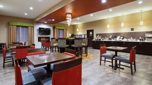A hotel dining area with multiple tables and chairs, a television, and a kitchen counter with food items. - Best Western Plus Fairview Inn & Suites (Fairview)