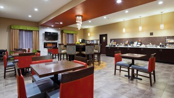A hotel dining area with multiple tables and chairs, a television, and a kitchen counter with food items.