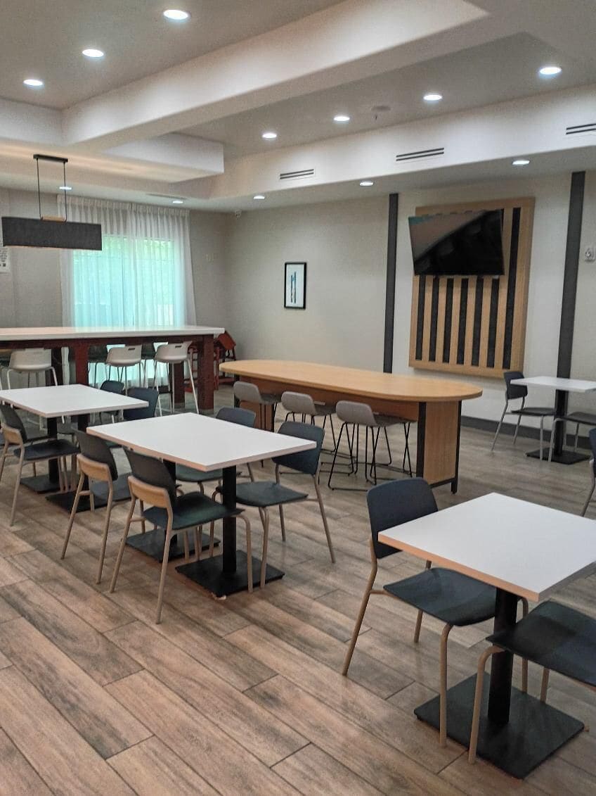 A modern indoor space with multiple tables and chairs, a large flat-screen TV mounted on the wall, and a wooden floor.