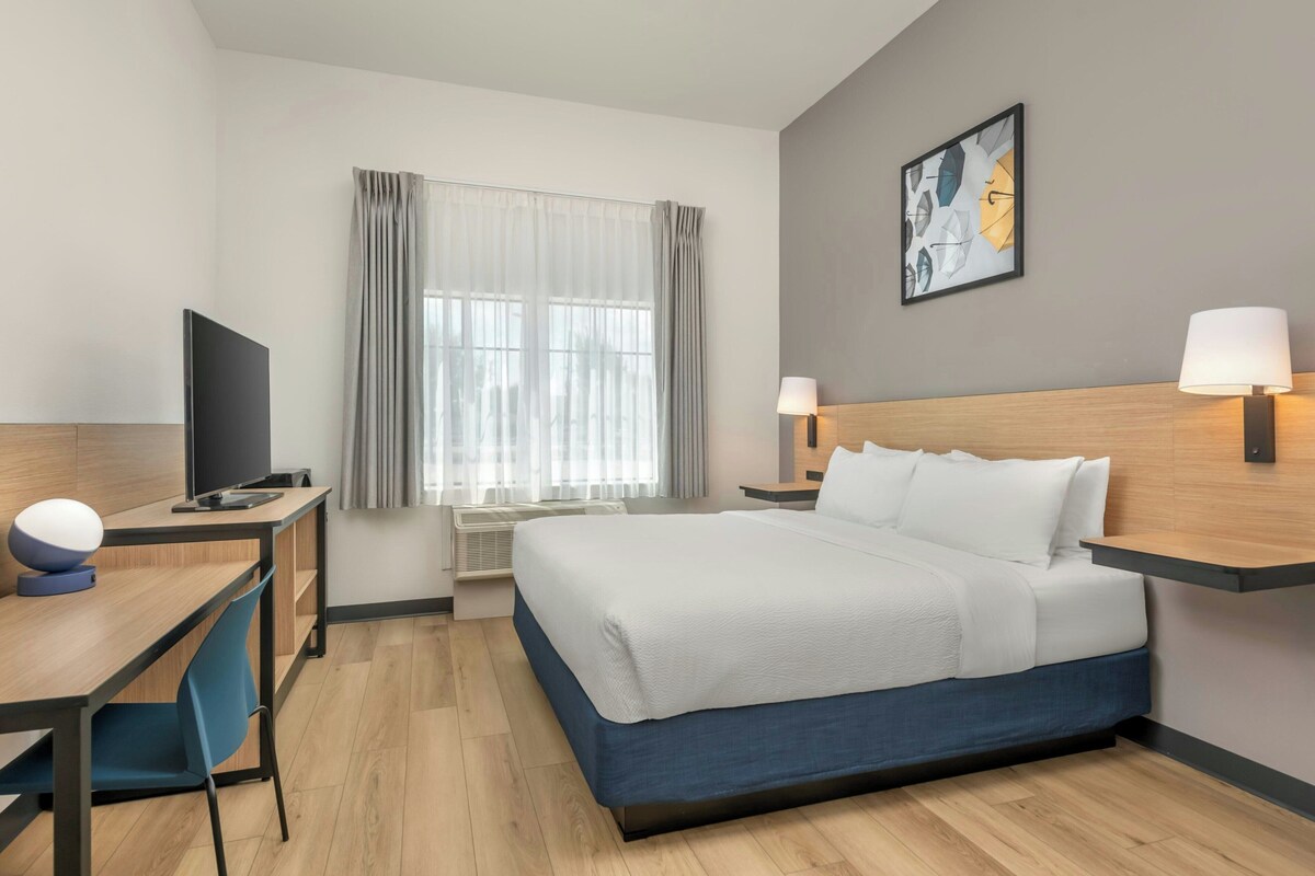 Habitación de hotel con cama, lámparas de noche, escritorio con silla, televisor y ventana con cortinas.