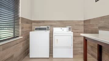 A laundry room with two white appliances, a window with blinds, and a wooden floor.