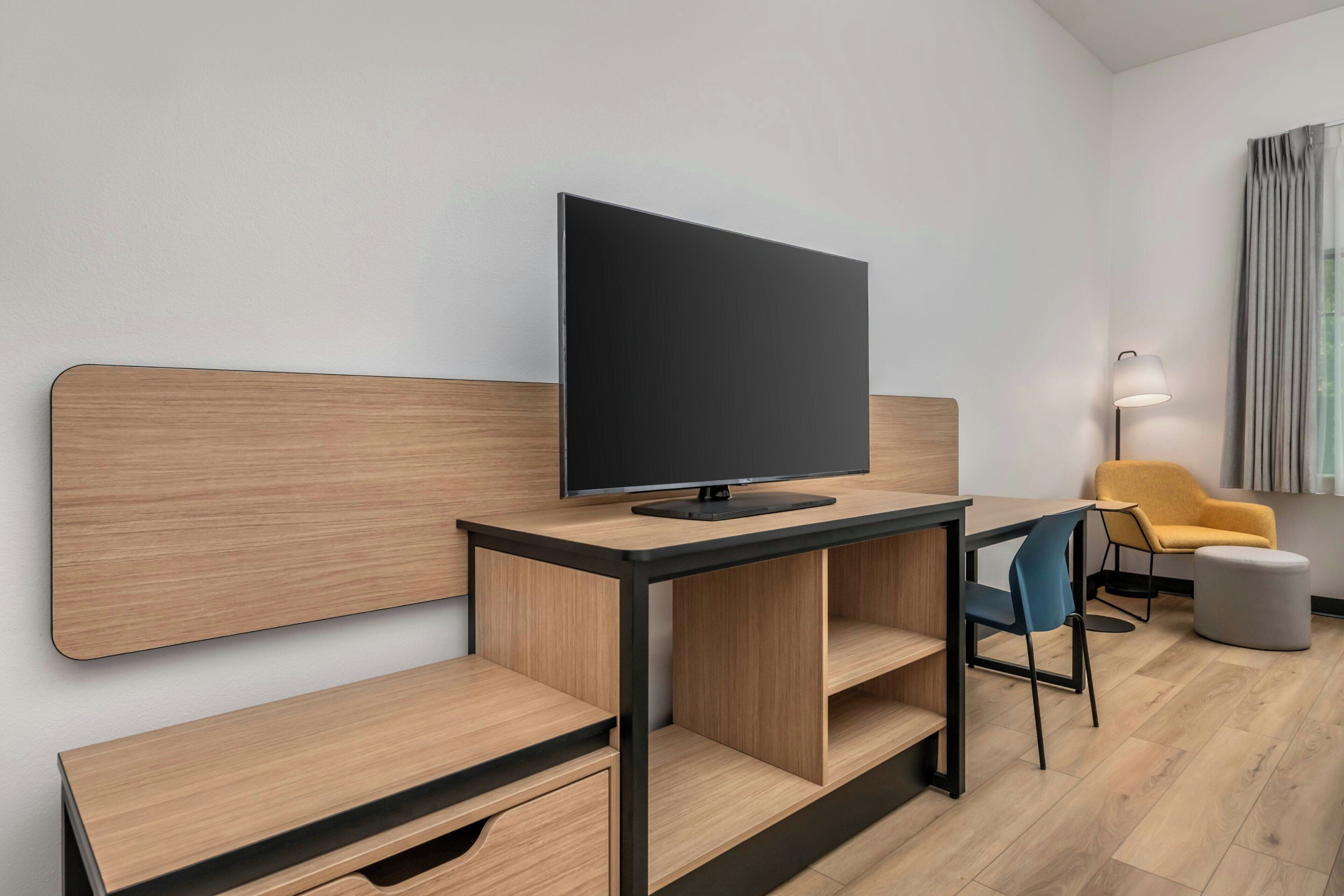 Une chambre d’hôtel moderne avec un bureau en bois, un téléviseur à écran plat, une chaise bleue, un fauteuil jaune et une lampe de chevet.