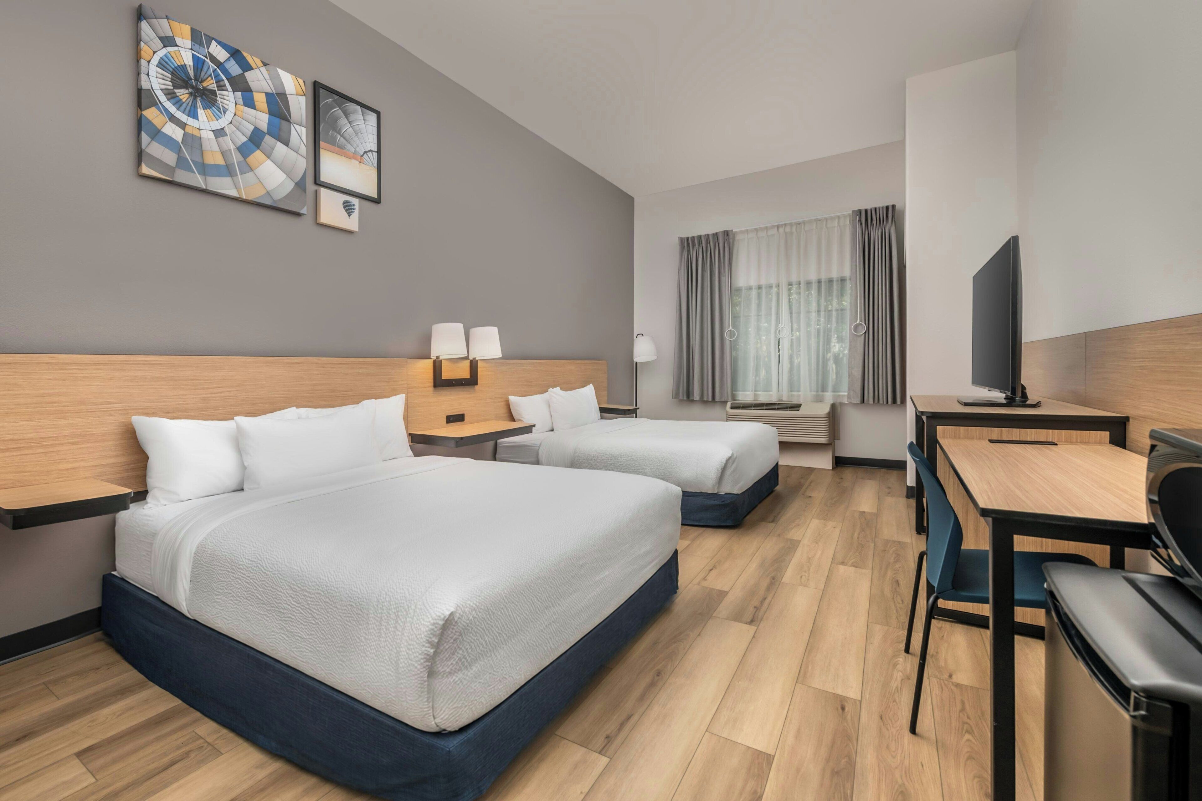 A hotel room with two beds, a desk, a chair, a TV, and artwork on the wall.