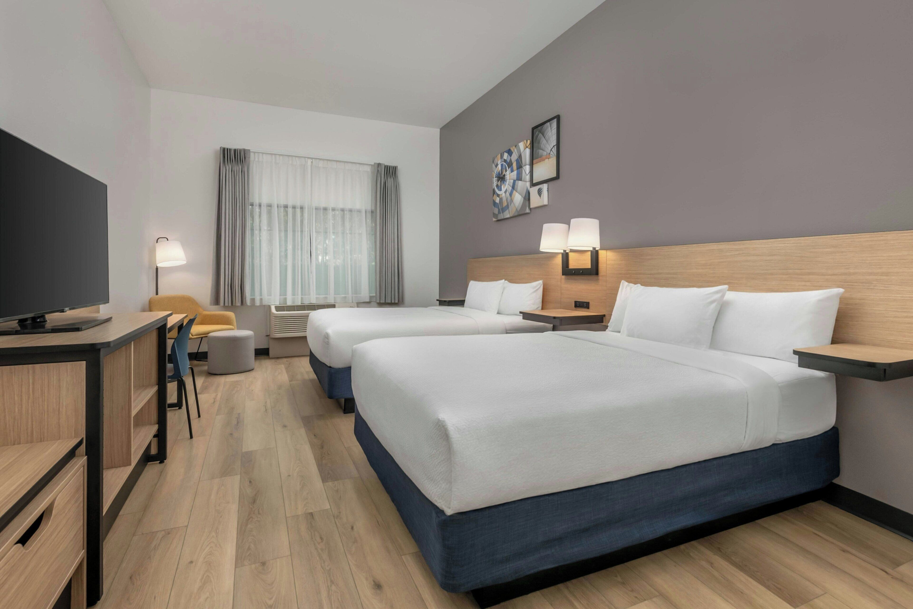Ein Hotelzimmer mit zwei Betten, einem Schreibtisch, einem Stuhl, einem Fernseher und einem Fenster mit Vorhängen.