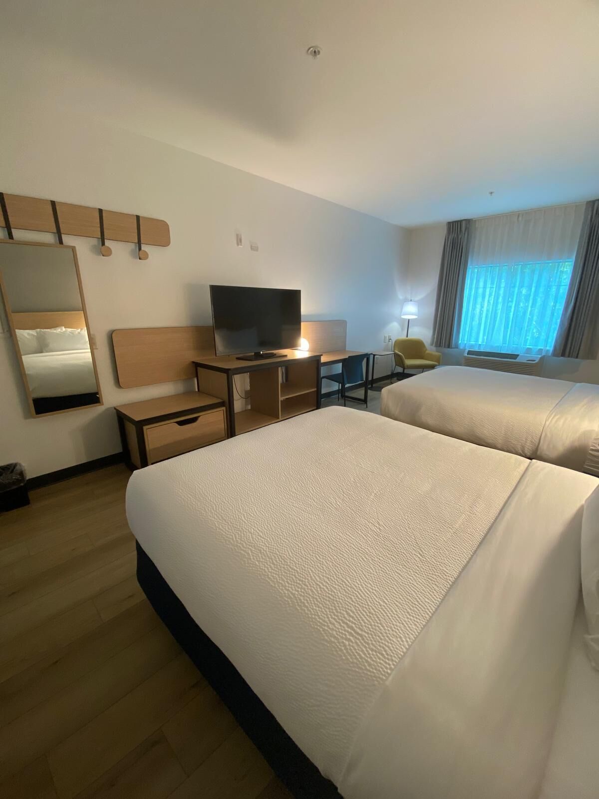 A hotel room with two beds, a desk with a TV, a chair, a mirror, and a window with curtains.