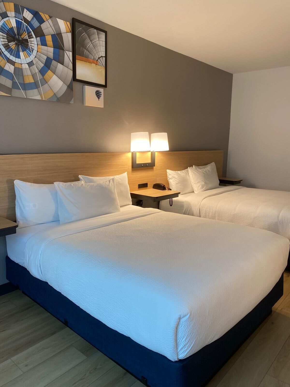 A hotel room with two beds, a headboard, bedside tables, lamps, and artwork on the wall.