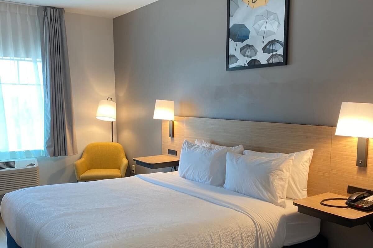 Habitación de hotel con una cama grande, dos lámparas de noche, una silla, un cuadro en la pared y una ventana con cortinas.