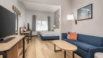 A modern hotel room with a flat-screen TV, a desk with a chair, a bed with a blue headboard, a blue sofa, and a small table.
