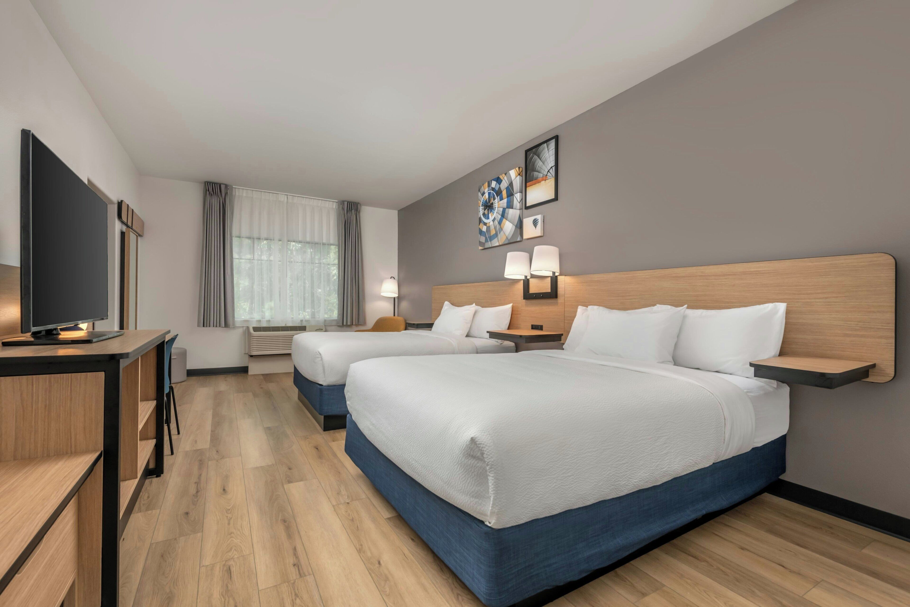 A hotel room with two beds, a desk with a flat-screen TV, a bedside table with lamps, and artwork on the wall.