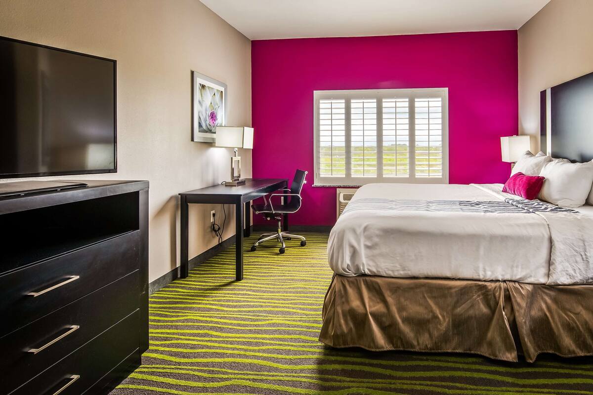A hotel room with a bed, a desk, a TV, and a window with blinds.