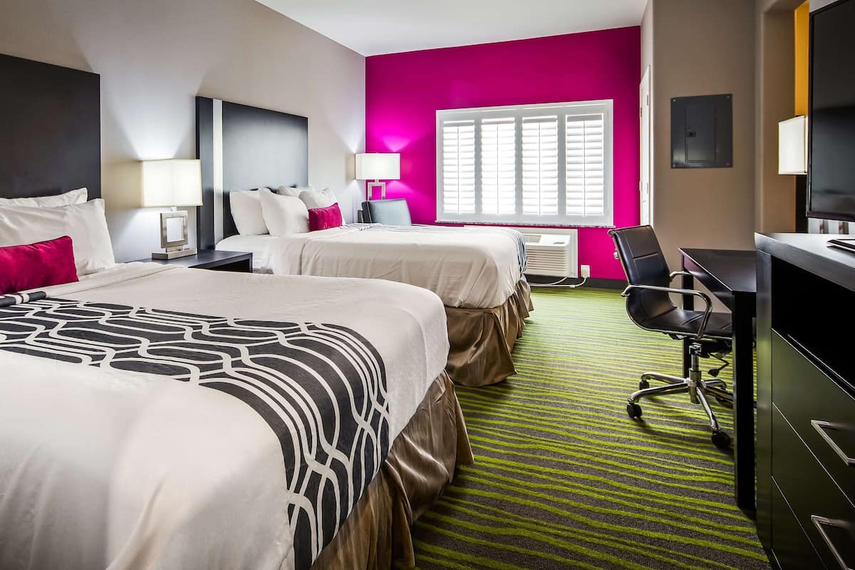 A hotel room with two beds, a desk, a chair, and a window with blinds.