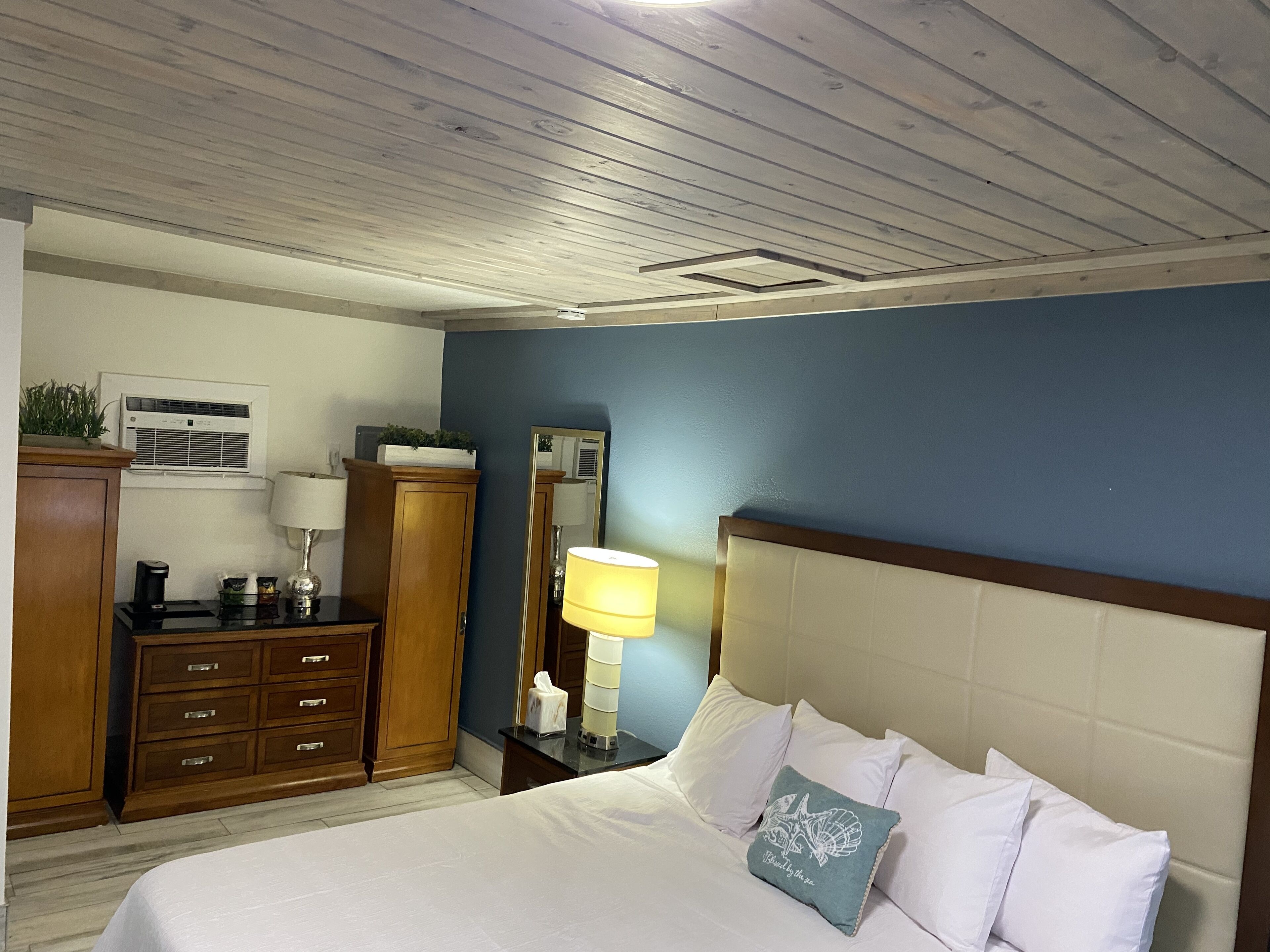 A bedroom with a bed, a nightstand, a dresser, a mirror, and a ceiling with wooden beams.