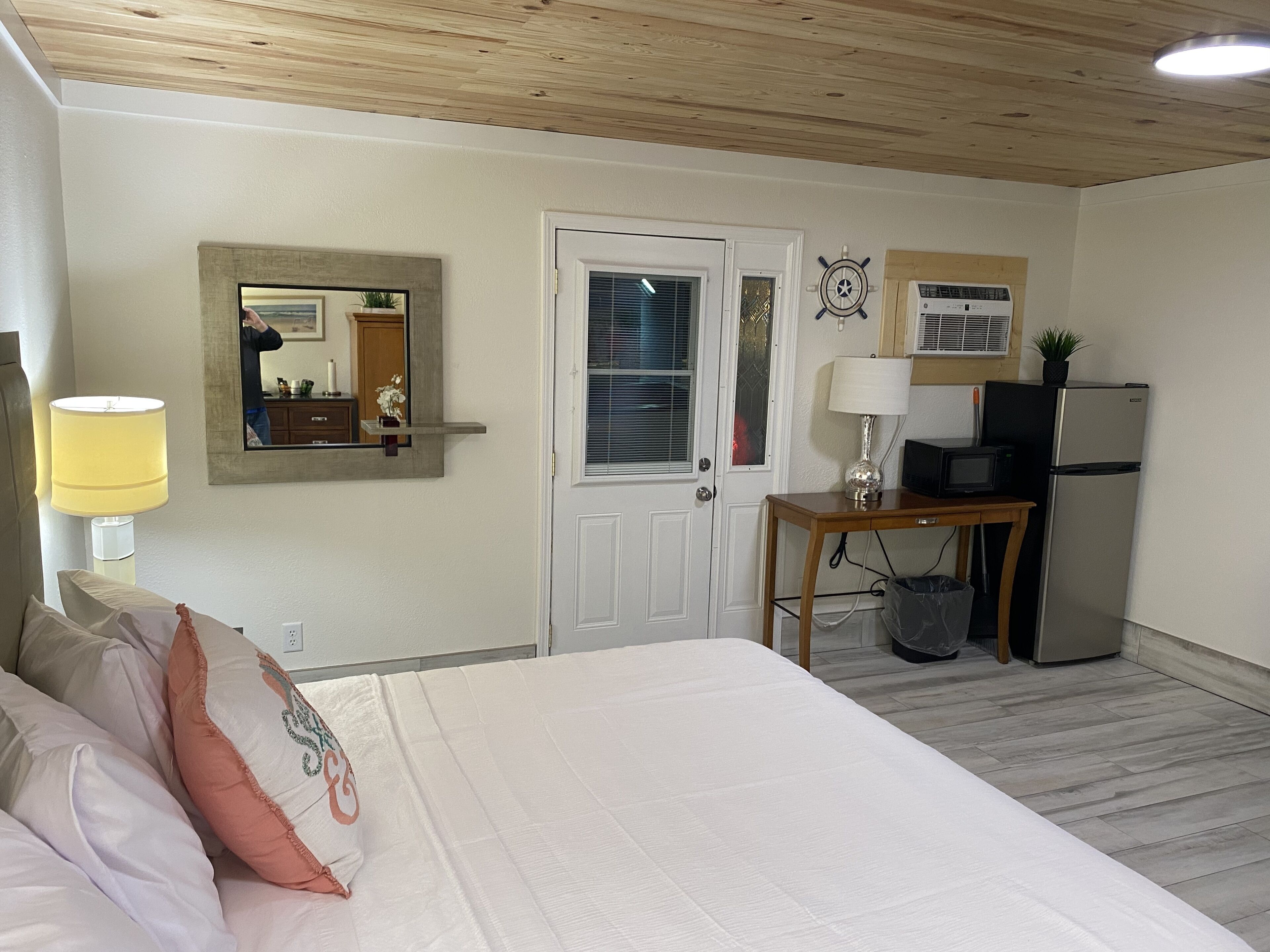 A bedroom with a bed, a desk with a lamp, a refrigerator, and a door with a window.