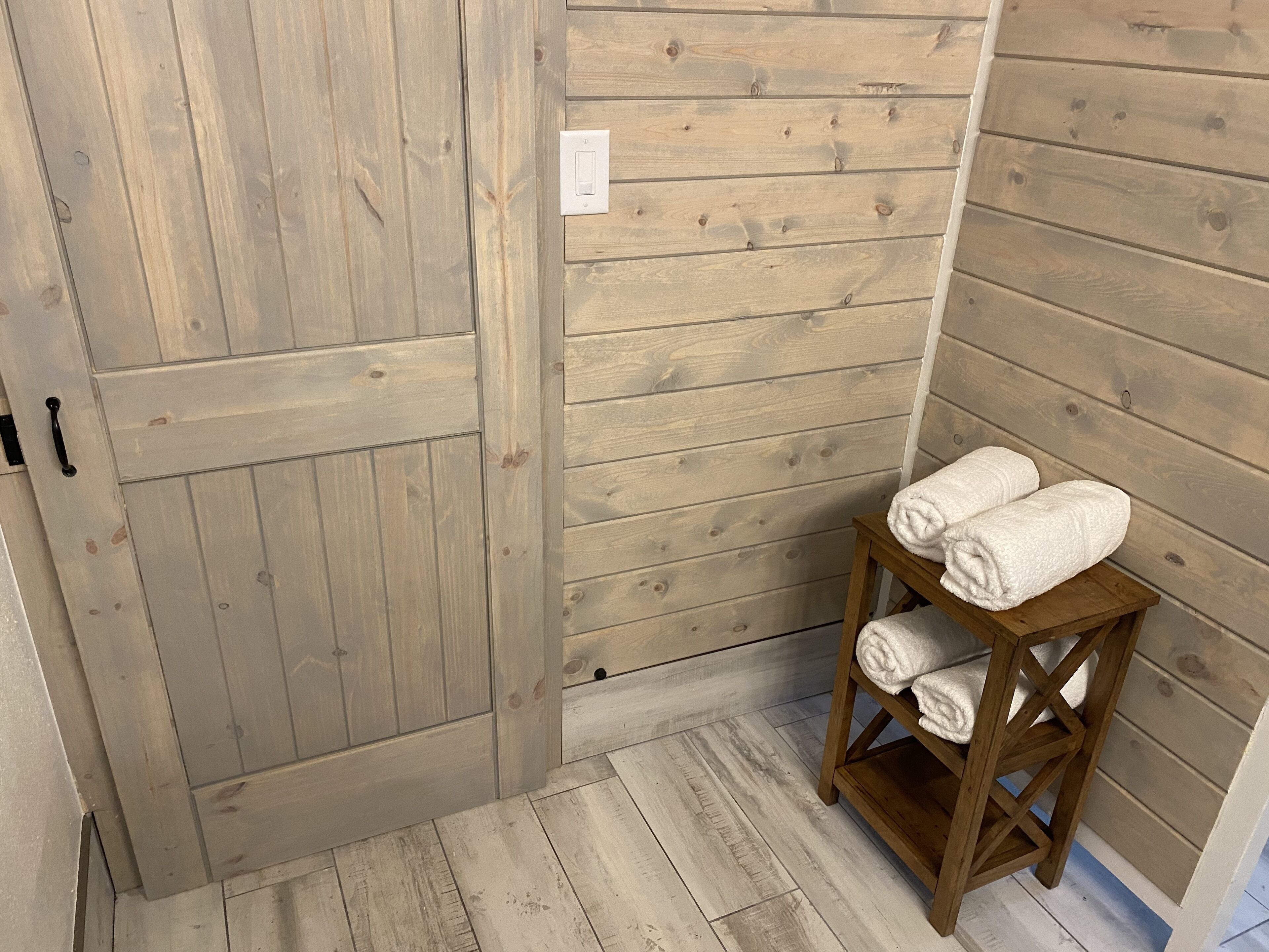 Une pièce rustique en bois, avec une grande porte en bois, un petit tabouret en bois sur lequel sont posées des serviettes, et un interrupteur sur le mur.