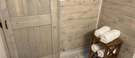 Une pièce rustique en bois, avec une grande porte en bois, un petit tabouret en bois sur lequel sont posées des serviettes, et un interrupteur sur le mur.