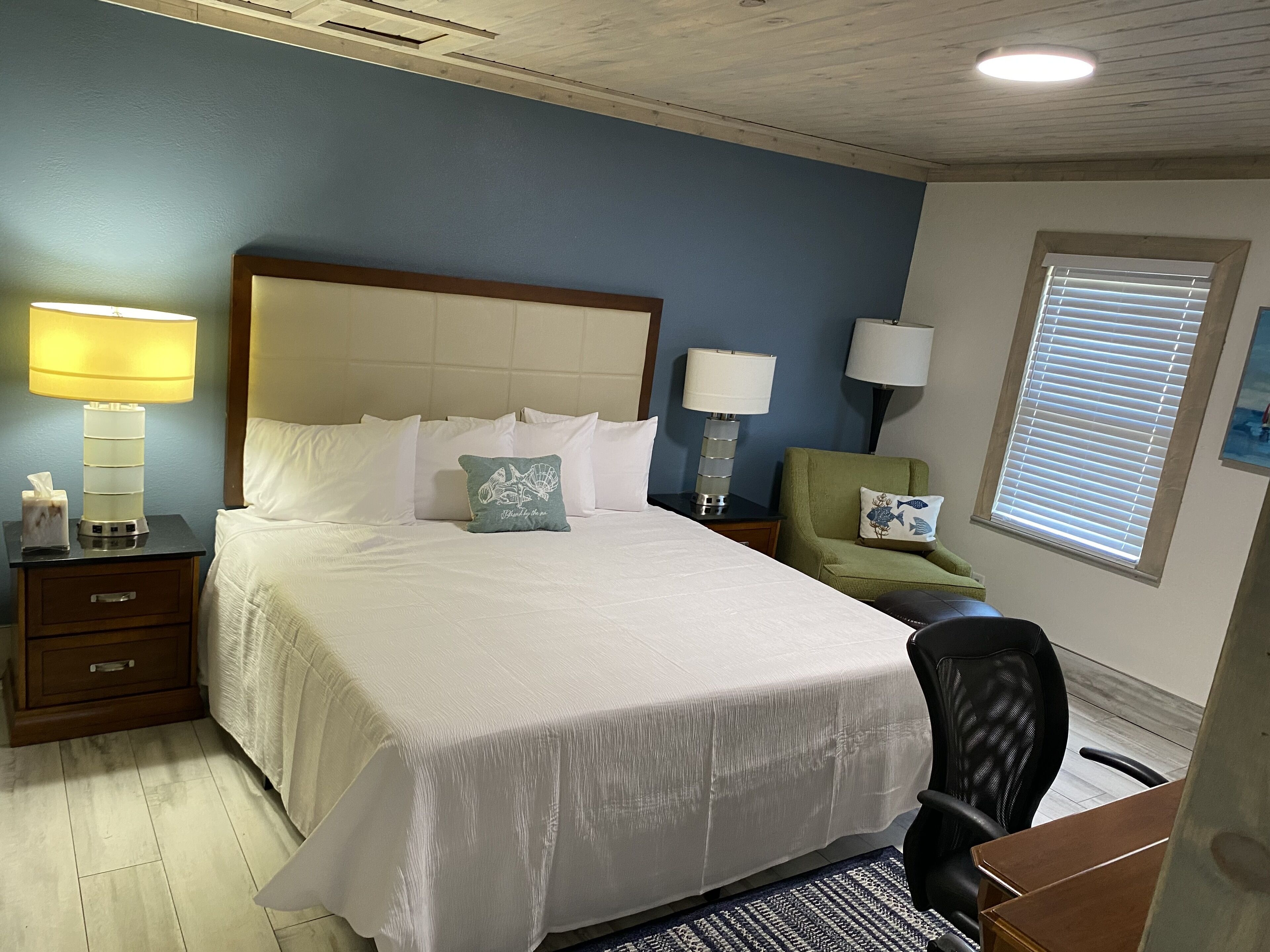A hotel room with a large bed, two bedside tables with lamps, a green armchair, a black office chair, and a window with blinds.