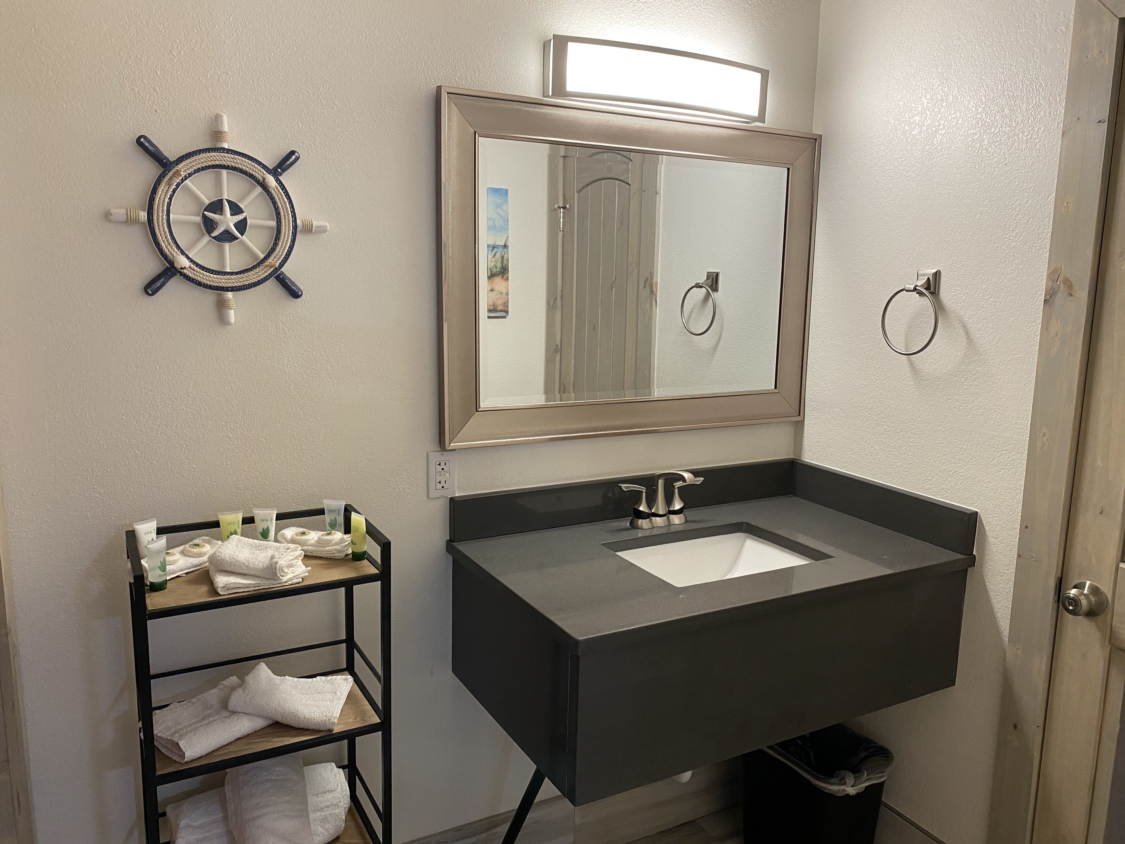 Une salle de bain dotée d’un grand miroir, d’une décoration en forme de roue de navire, d’un porte-serviettes et d’un lavabo avec un plan de travail noir.