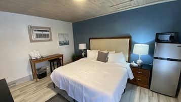 A hotel room with a bed, bedside table, lamp, refrigerator, and a desk with a chair.