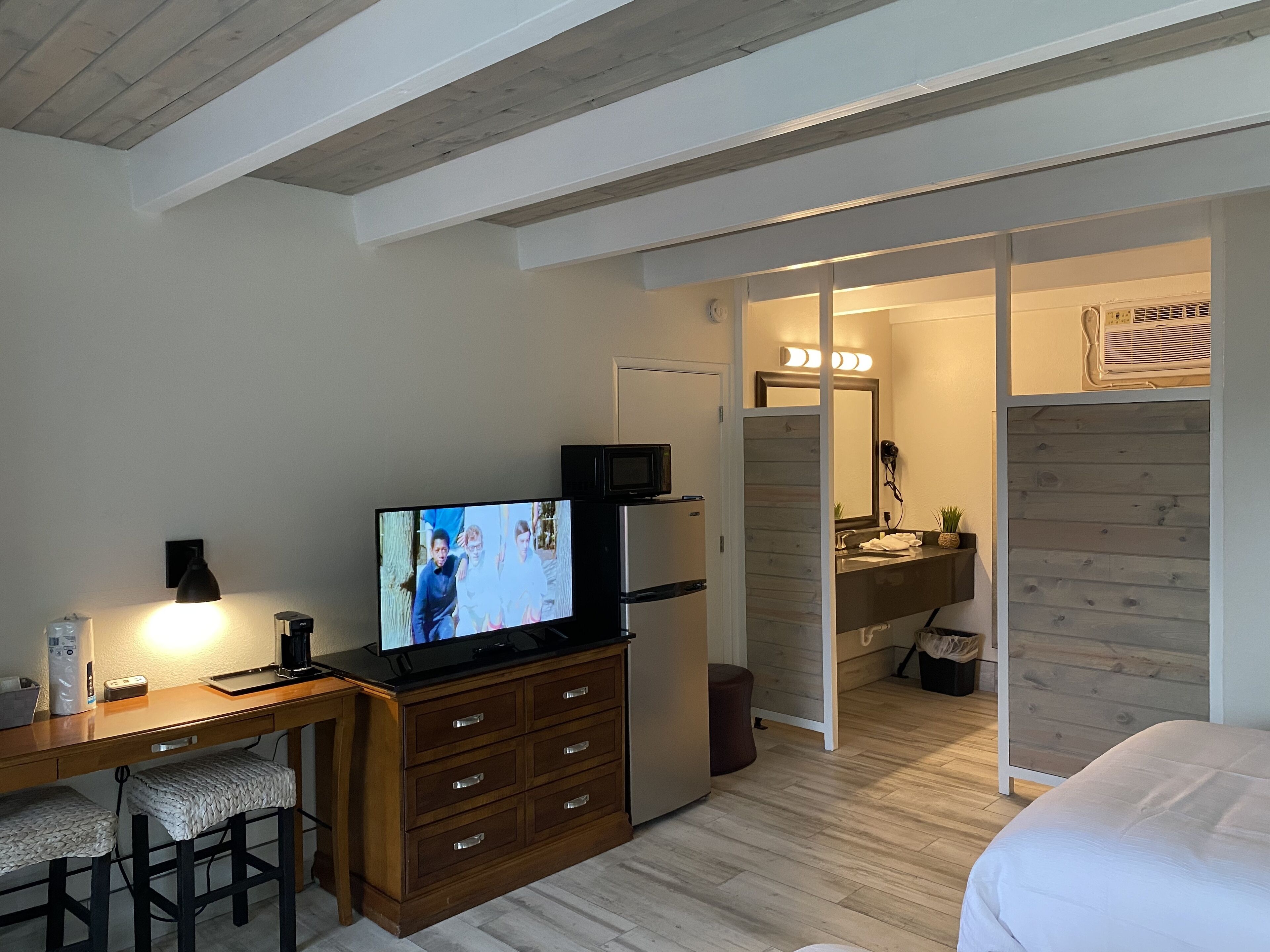 Ein Hotelzimmer mit Holzschreibtisch, Fernseher, Bett und Badezimmer mit Holztischchen und Spiegel.
