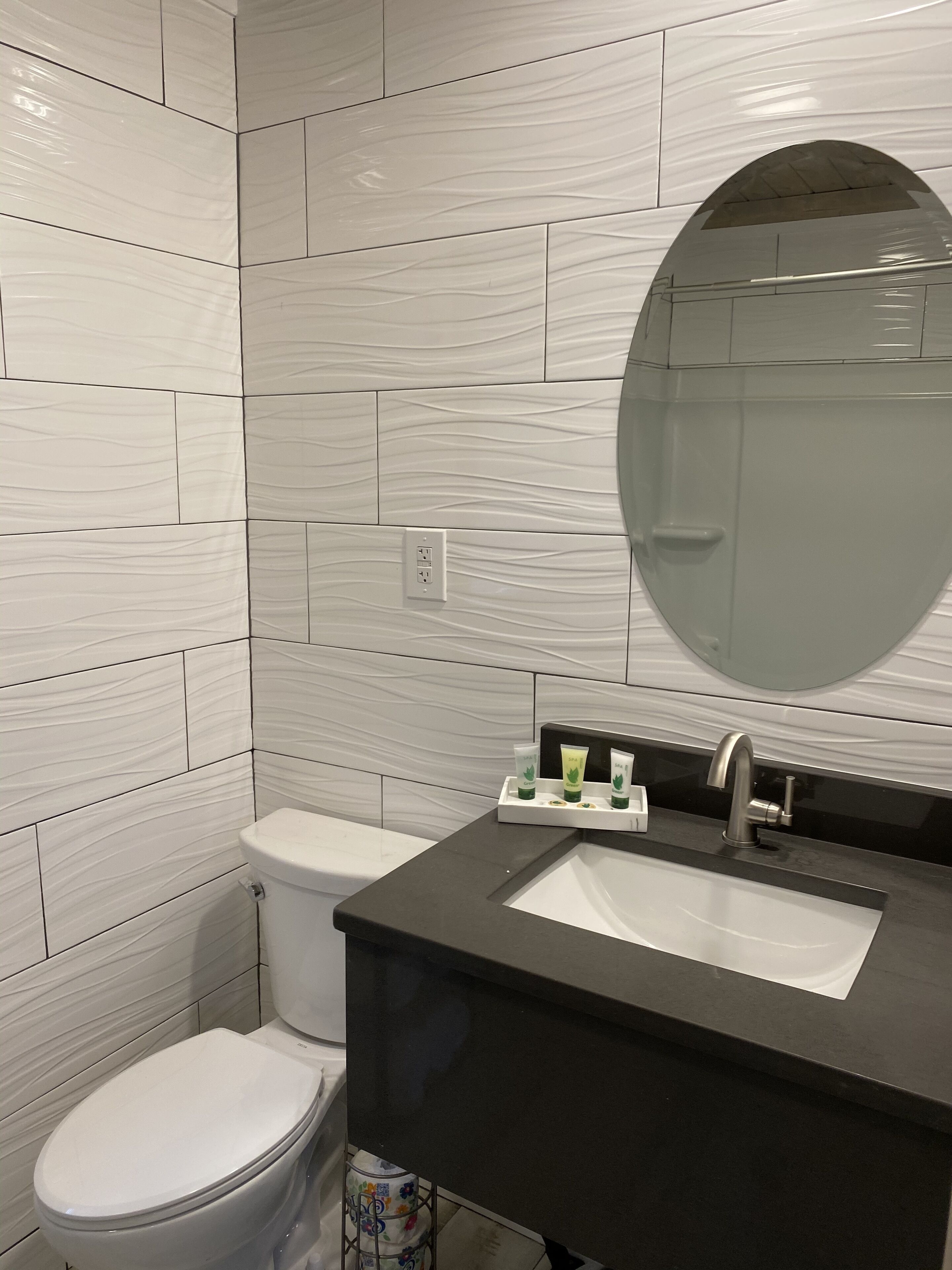 Une salle de bain avec des carreaux de marbre blanc, un lavabo noir, un miroir et des articles de toilette.