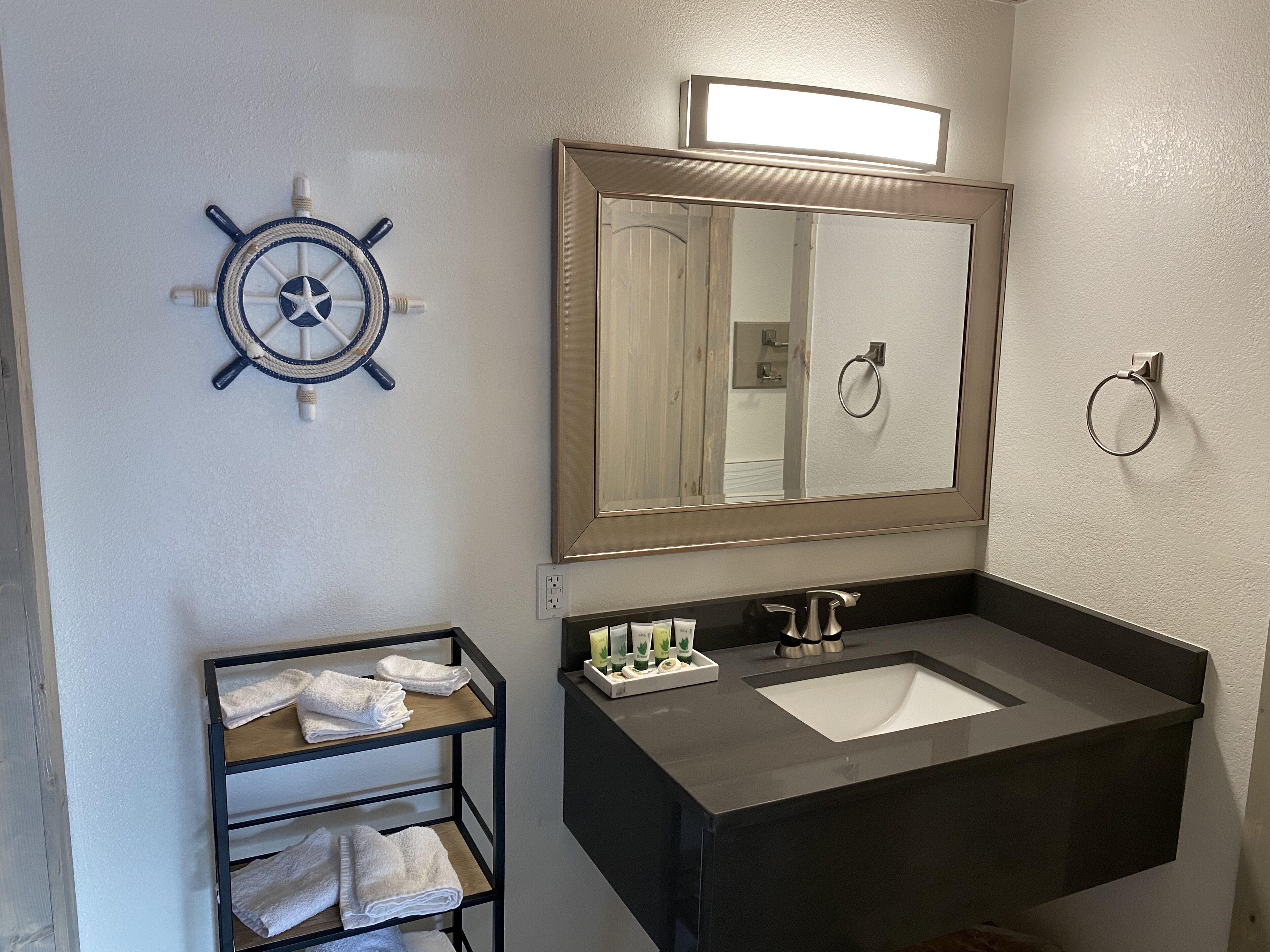 A bathroom with a dark sink, a large mirror, a ship's wheel decoration, and a towel rack with folded towels.