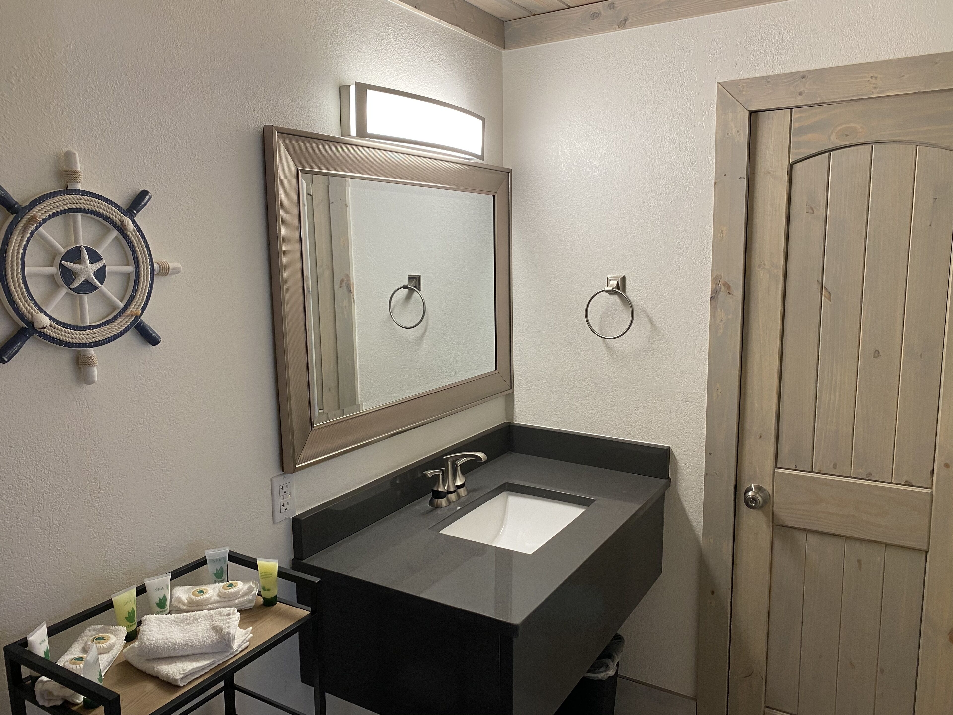 Une salle de bain avec un lavabo noir, un miroir, un élément décoratif en forme de roue de navire et une porte en bois.