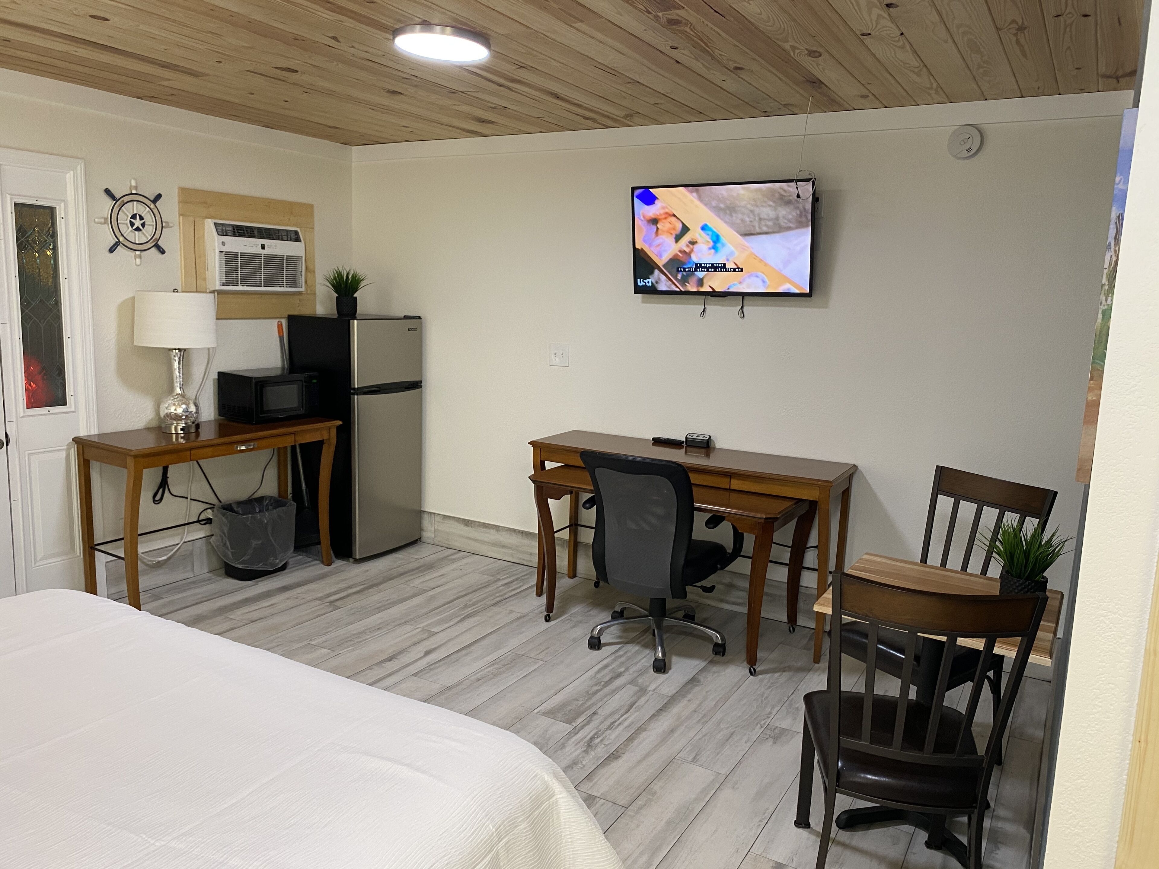 A hotel room with a bed, a desk with a computer, a TV, a refrigerator, and a small trash bin.