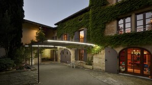 Un patio con zona de estar cubierta, muros de piedra y un edificio con ventanas y puertas de arco.