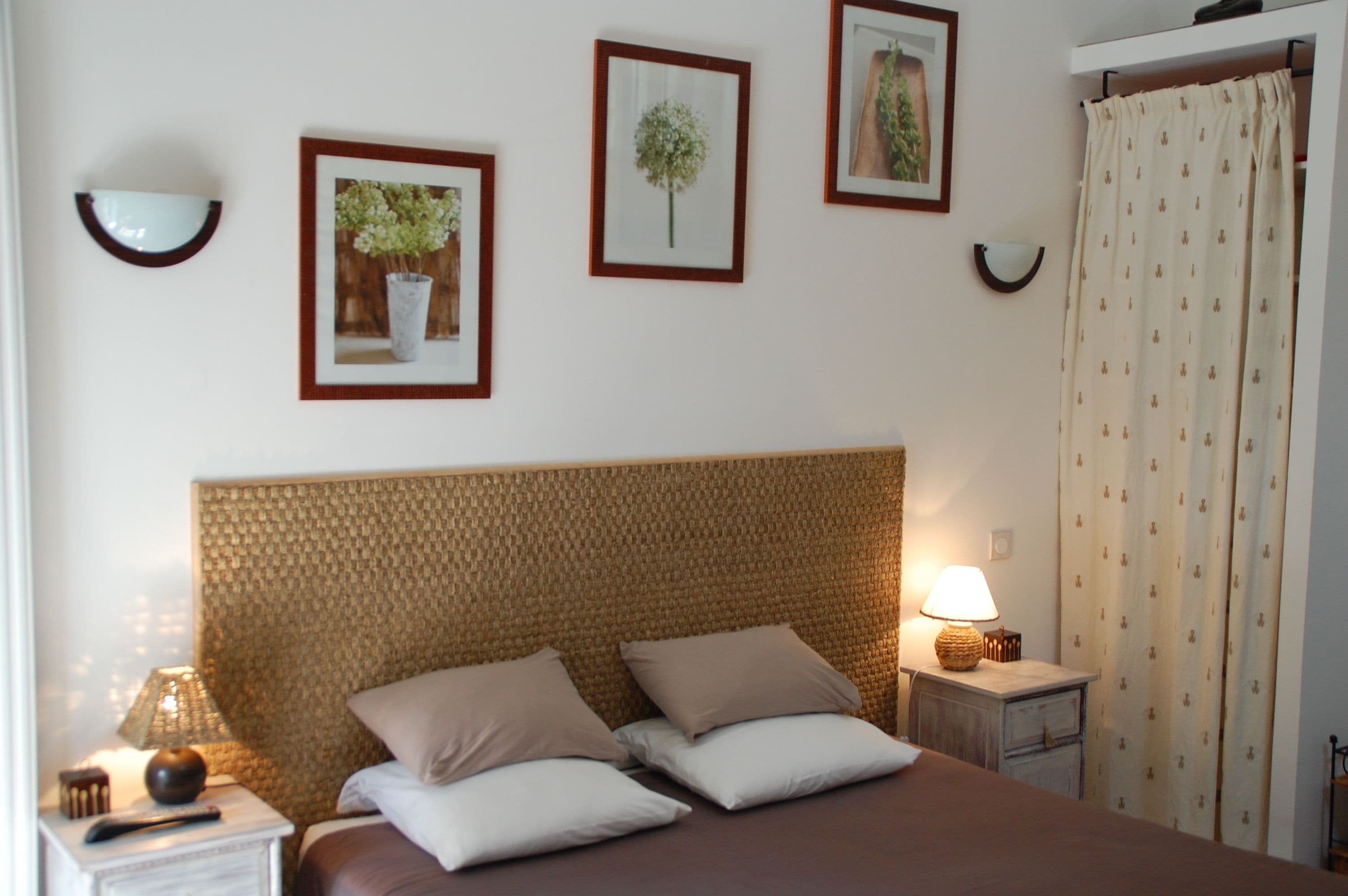 Quarto com cabeceira de fibra natural, duas camas com roupa de cama bege e branca, mesa de cabeceira com abajur e quadros decorativos na parede.