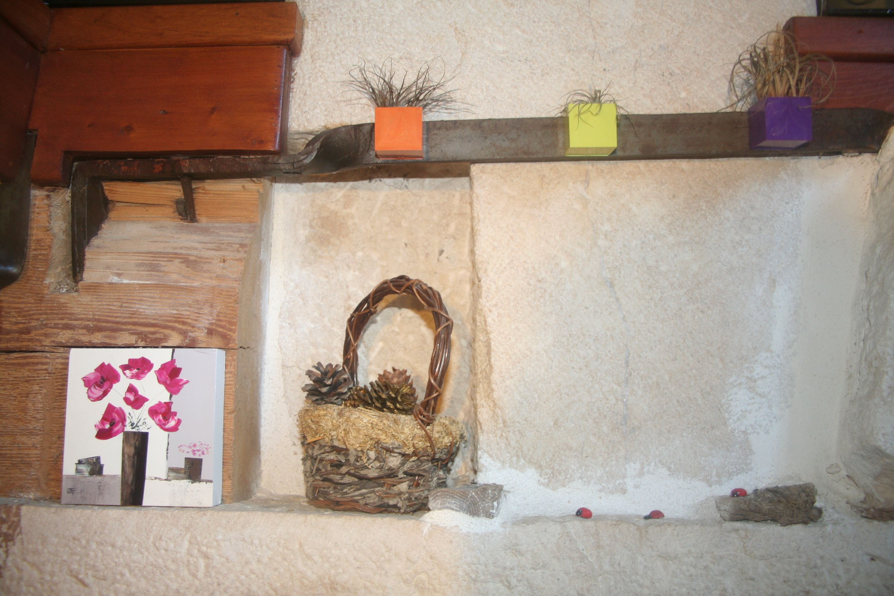 Un interior rústico con una repisa de madera que sostiene objetos decorativos, una cesta tejida con piñas y un cuadro enmarcado.