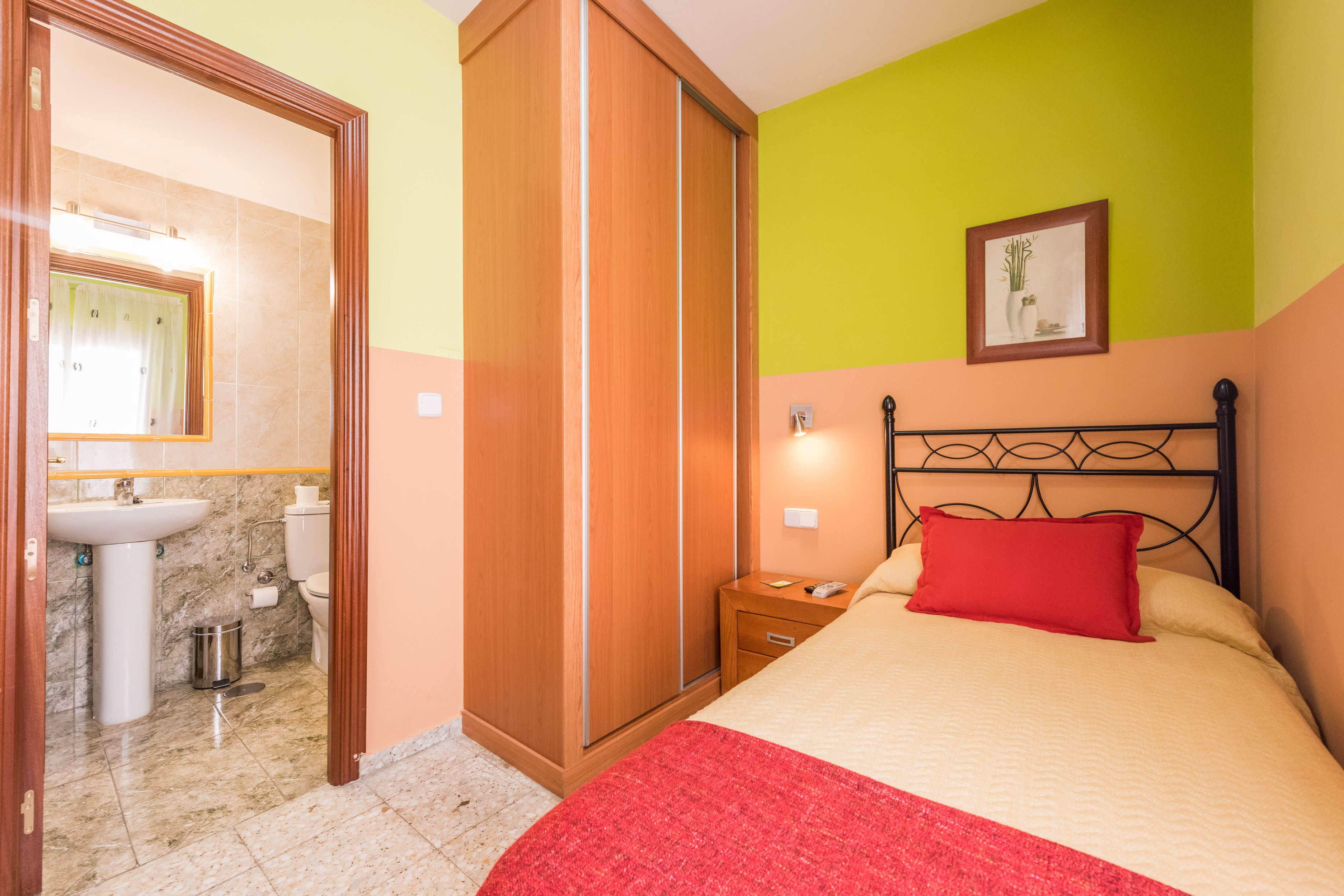 Quarto com cama, mesa de cabeceira, travesseiro vermelho, guarda-roupa de madeira, banheiro com pia e vaso sanitário, e uma moldura na parede.