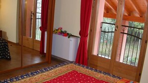 A bedroom with a large mirror, a red and blue patterned rug, a white radiator, and a view of greenery through a barred window.