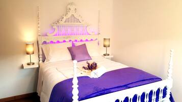 Una cama con cabecera blanca, dos lámparas de noche y una colcha morada.
