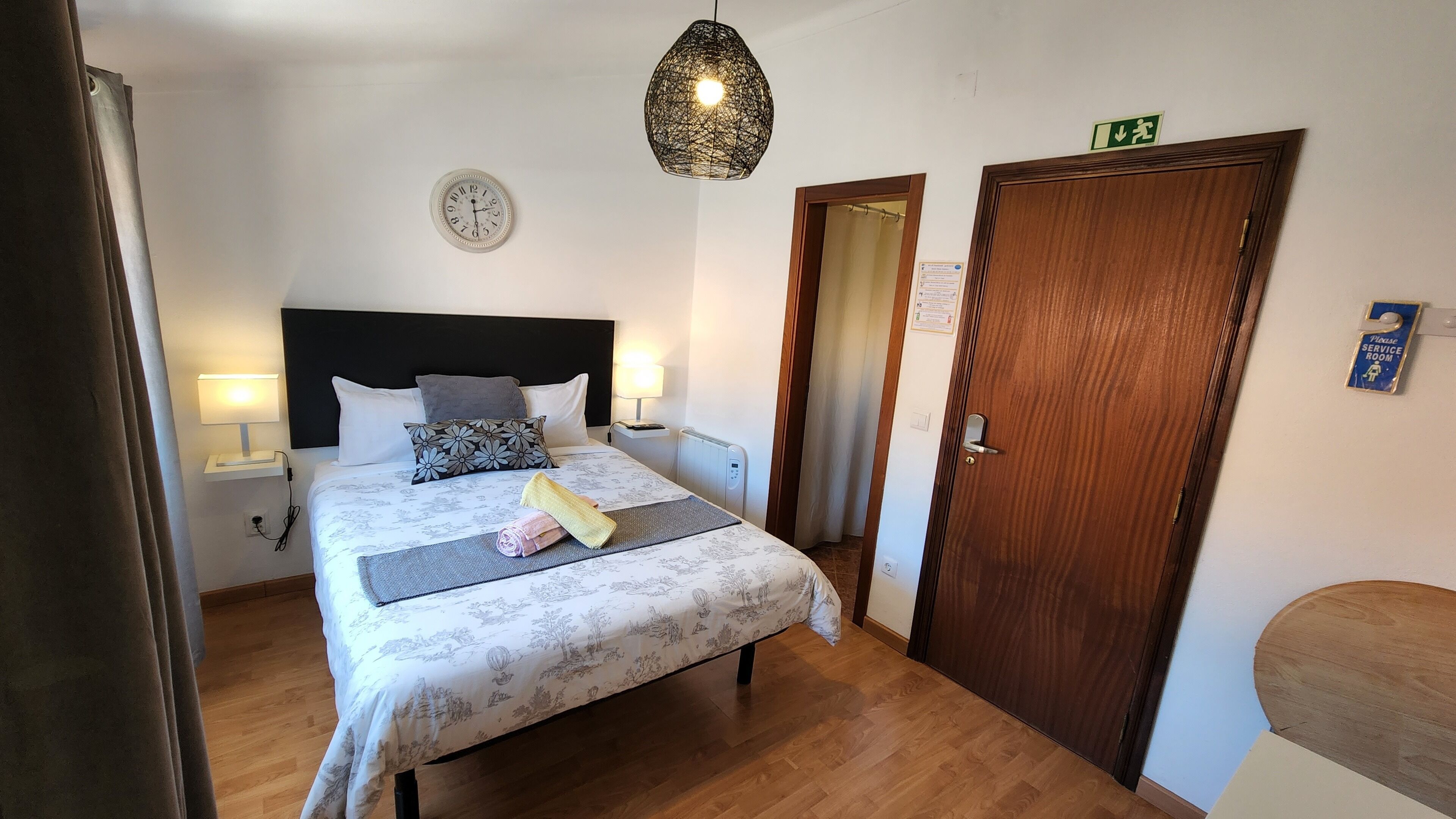 A hotel room with a bed, bedside lamps, a clock, a door, and a sign indicating a service room.