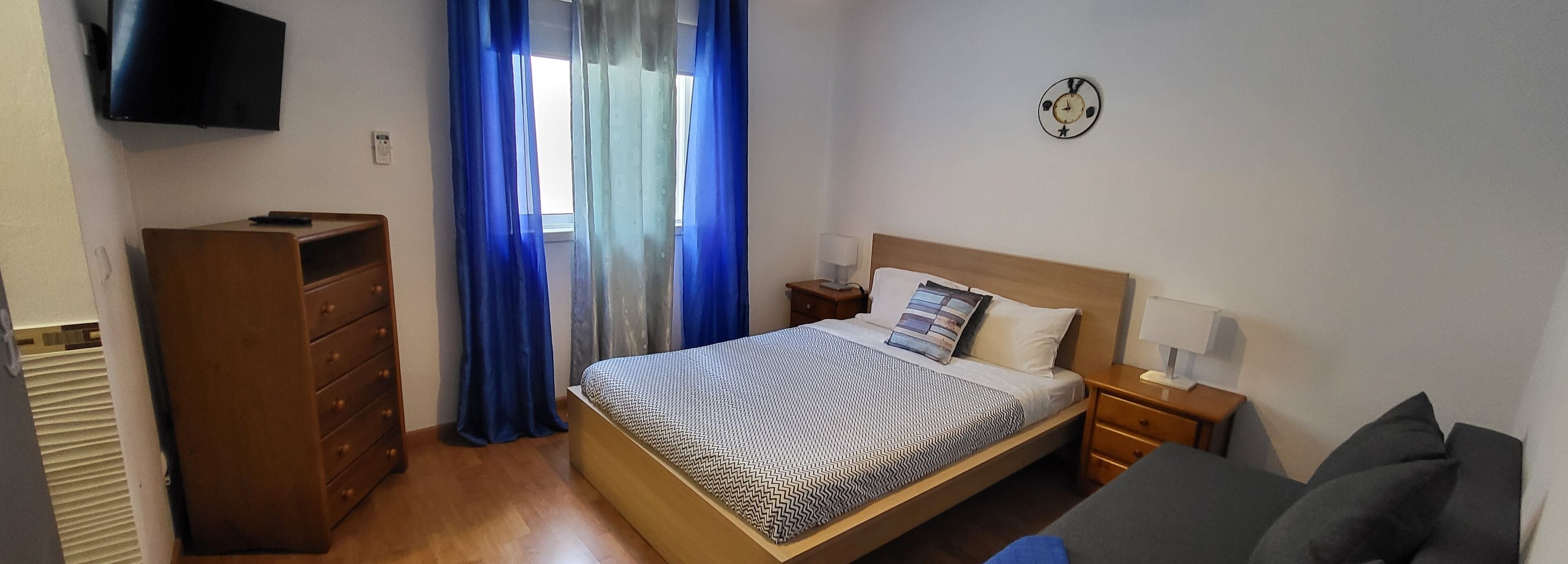 A bedroom with a bed, a dresser, a TV, a clock, and a window with blue curtains.