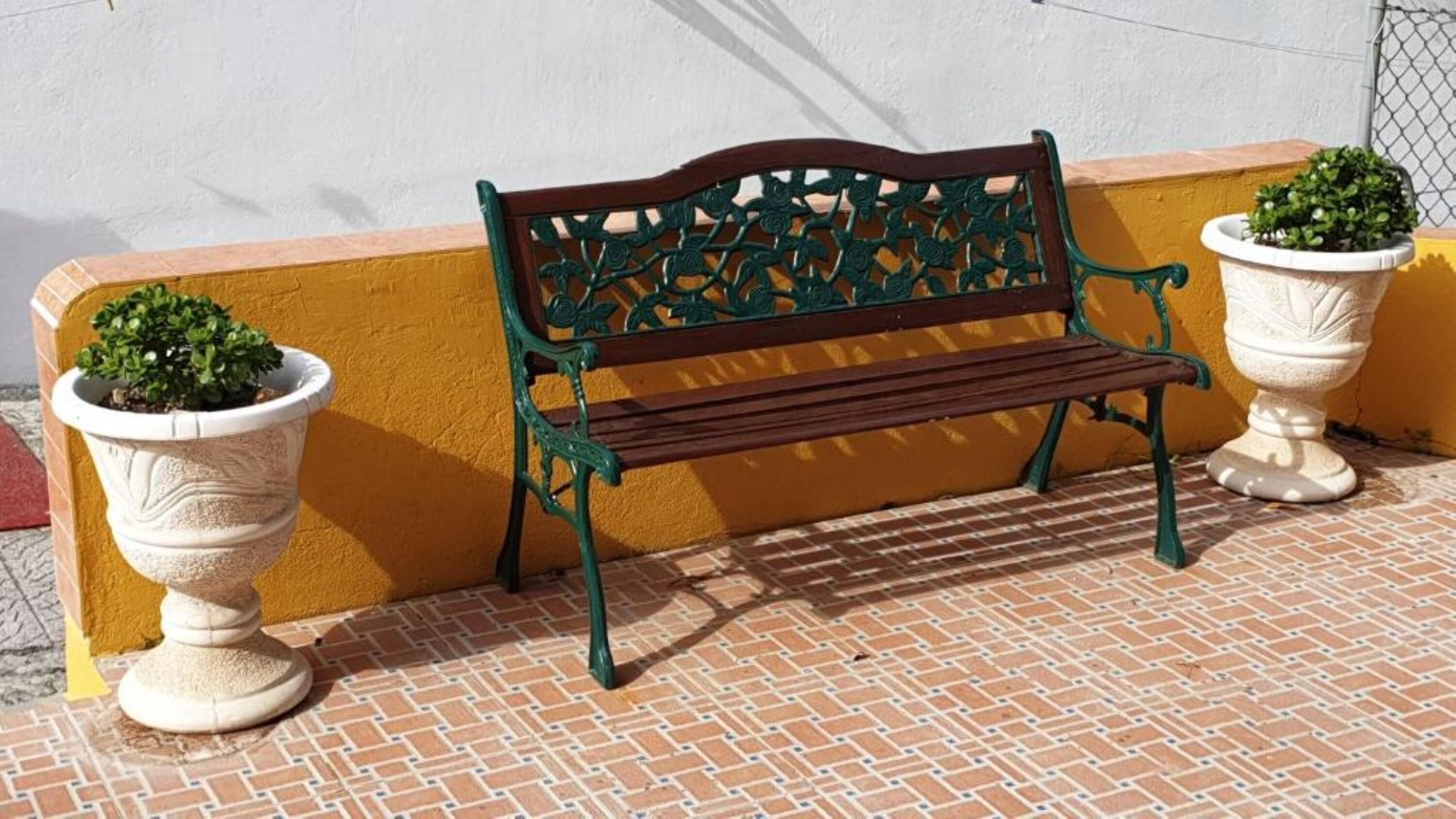 Un banco con ornamentación metálica elaborada, situado entre dos macetas blancas grandes con plantas verdes.