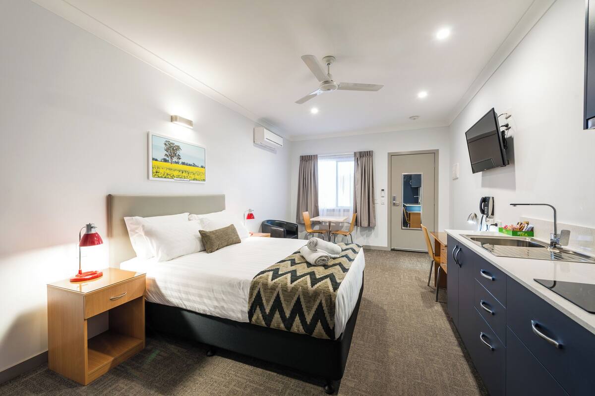 Ein Hotelzimmer mit Bett, Nachttisch, Lampe, Fernseher und einer Küchenzeile.