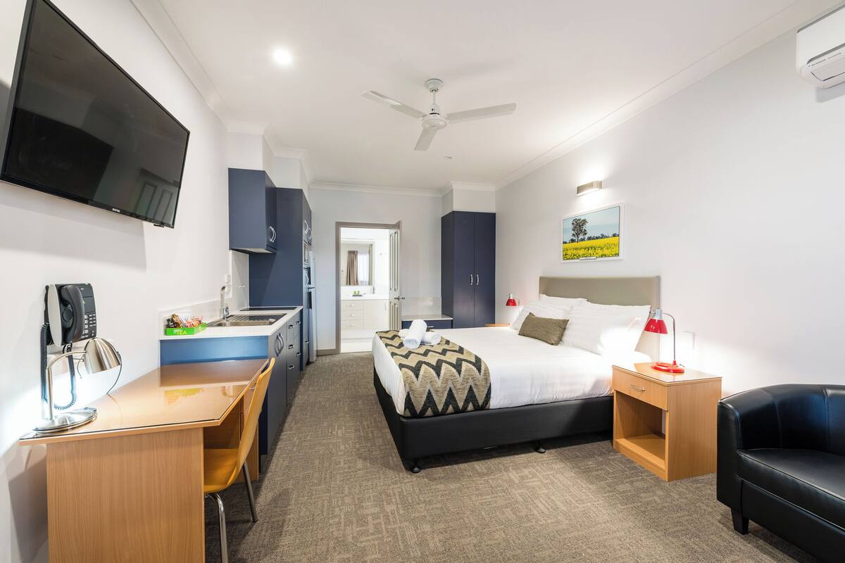 Ein Hotelzimmer mit Bett, Schreibtisch, Sessel, Fernseher, Deckenventilator und Bildern an der Wand.