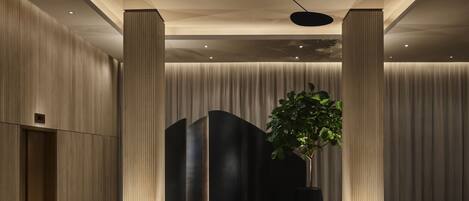 A modern lobby with a central seating area, a large potted plant, and a ceiling with unique lighting fixtures.