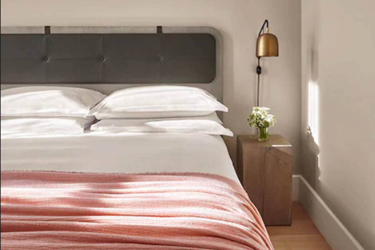 Een net opgemaakt bed met witte lakens, een grijs hoofdbord en een gestreept rode deken. Een klein nachtkastje met een lamp en een vaas bloemen is zichtbaar.