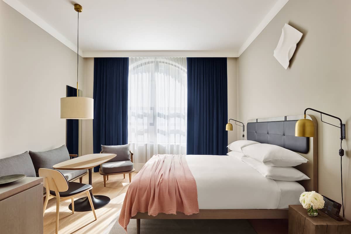 Een hotelkamer met een bed, nachtkastje, stoel, bureau, lamp en een kleine tafel met een bloevas.