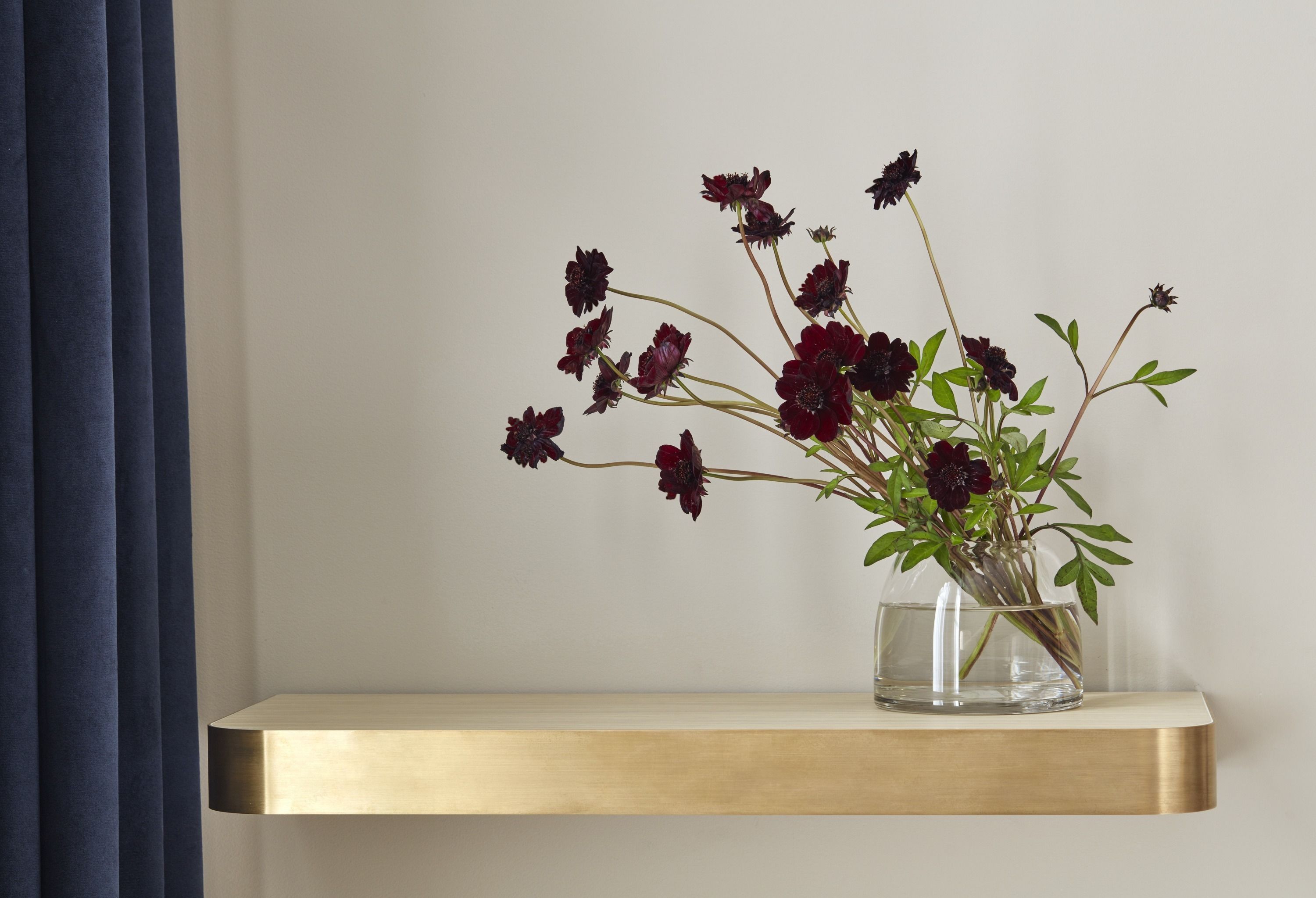 A clear glass vase with water and dark red flowers on a wooden shelf against a plain wall.