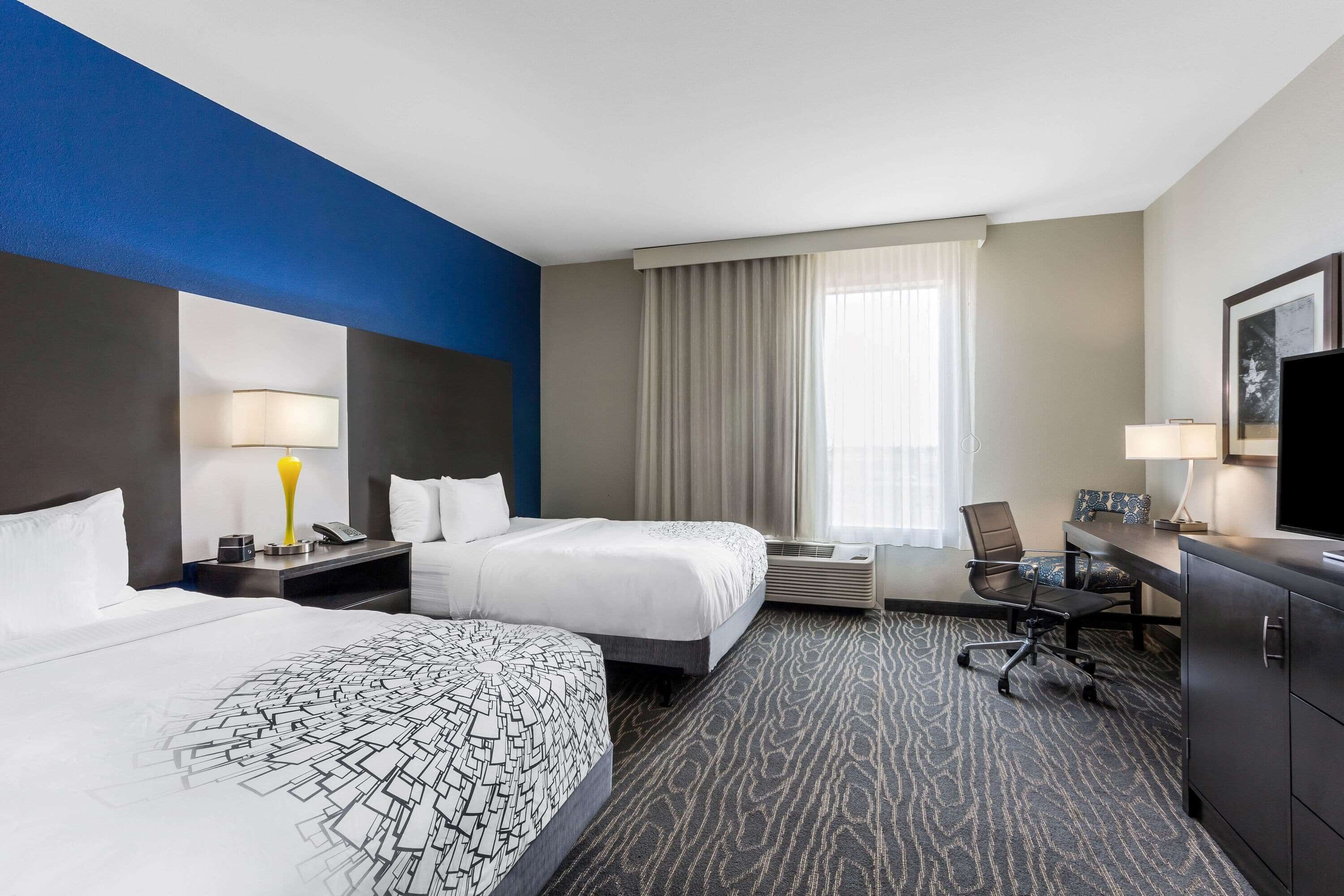 A hotel room with two beds, a desk, a chair, a TV, and a large window with curtains.