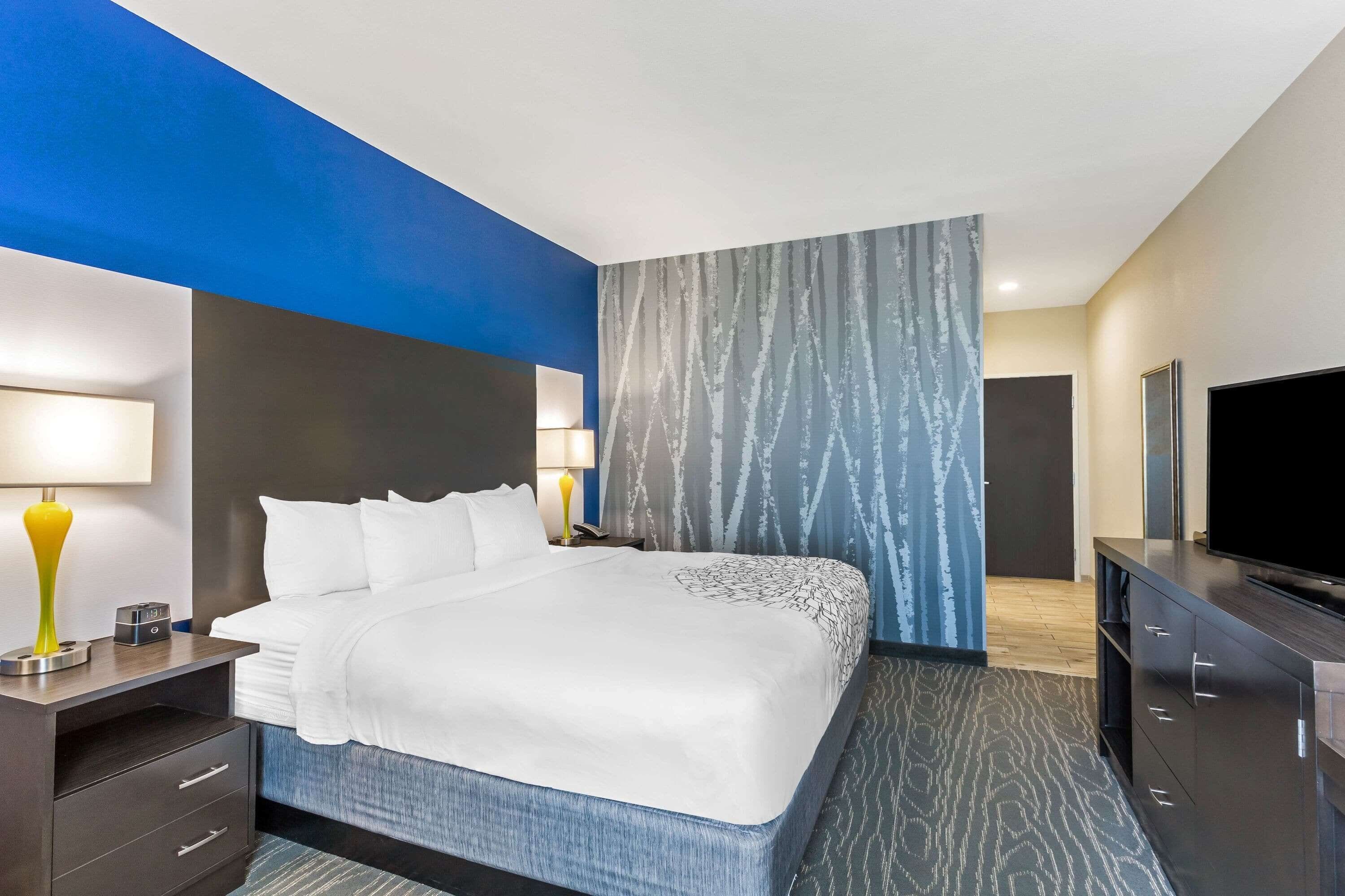 A modern hotel room with a large bed, bedside tables, a flat-screen TV, and a patterned wall.