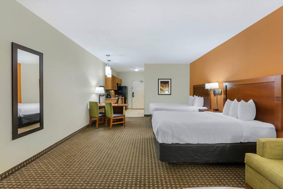 A hotel room with two beds, a desk with a chair, a mirror, and a kitchenette.